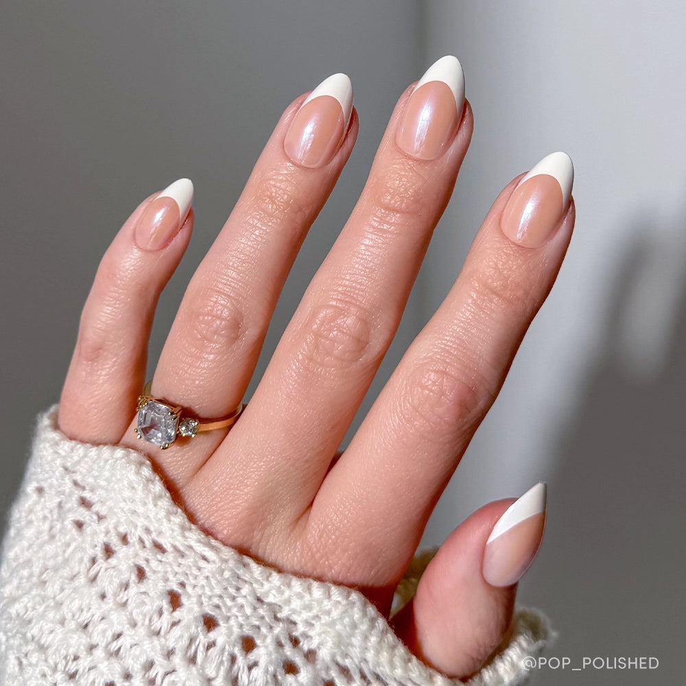A hand with short almond nails sports glamnetic Mrs. press-ons in a modern French tip design, complemented by a gold gemstone ring and a textured white knit sleeve—perfect bridal nail inspo.